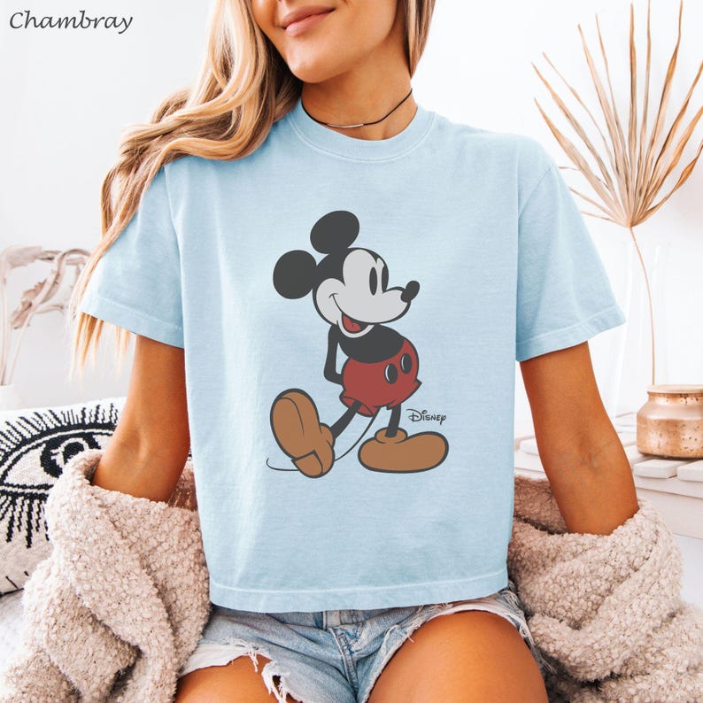 以下が含まれることがあります： ミッキーマウスのグラフィックが特徴のライトブルーのTシャツ。漫画のキャラクターは、象徴的な赤いショートパンツ、茶色の靴、黒い耳で描かれています。ディズニーのロゴが見えます。柔らかい素材で作られたシャツです。