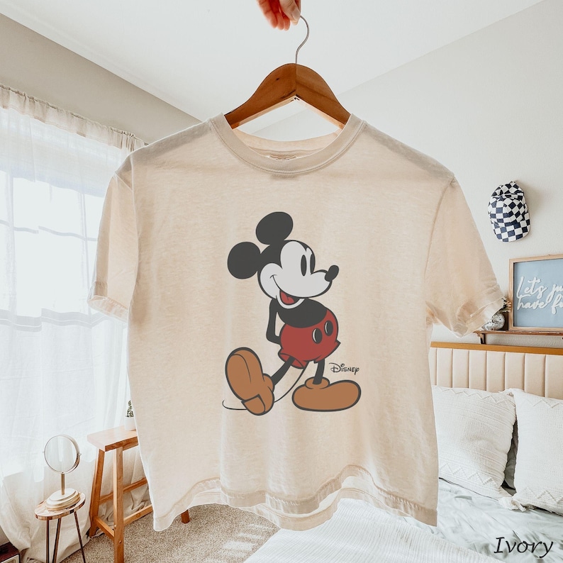 May include: Ivory-coloured t-shirt featuring a classic Mickey Mouse graphic. The cartoon character is depicted in black, red, and brown, with the Disney logo below. The shirt is hanging on a wooden hanger.