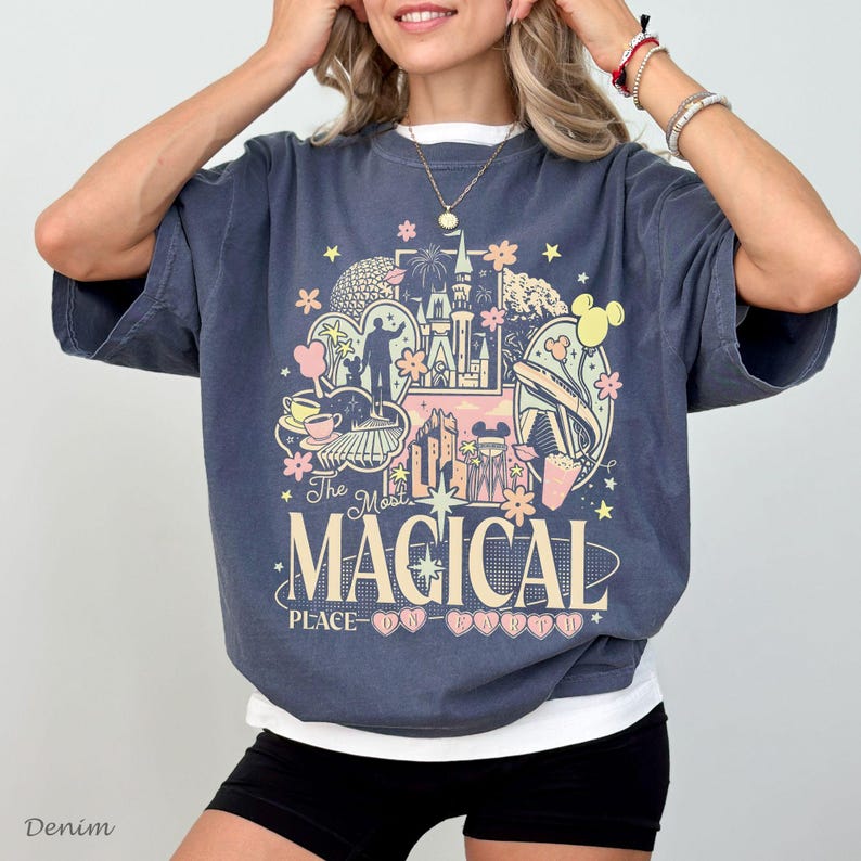 May include: A charcoal gray short-sleeve t-shirt featuring a multi-colored graphic print of a castle, park icons, and flowers. The text reads The Most Magical Place On Earth in a large serif font, layered over a white crew neck undershirt.