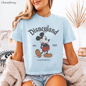 May include: Light blue t-shirt featuring a classic Disneyland graphic. The design includes the word "Disneyland" in a stylized font, Mickey Mouse, and the text "est. 1955 CALIFORNIA". The shirt has short sleeves and a crew neck.