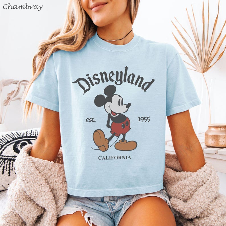 May include: Light blue t-shirt featuring a classic Disneyland graphic. The design includes the word "Disneyland" in a stylized font, Mickey Mouse, and the text "est. 1955 CALIFORNIA". The shirt has short sleeves and a crew neck.