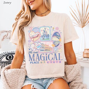 May include: Ivory-colored t-shirt featuring a colorful graphic design with the text "The most MAGICAL PLACE ON EARTH." The design includes illustrations of a castle, train, and other theme park elements. The shirt is short-sleeved and has a crew neck.