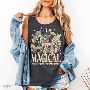 May include: A charcoal grey sleeveless tank top featuring a graphic print of a castle, a monorail, and various park icons in pastel pink, yellow, and white. The text reads The Most Magical Place on Earth, with small heart icons below the text.