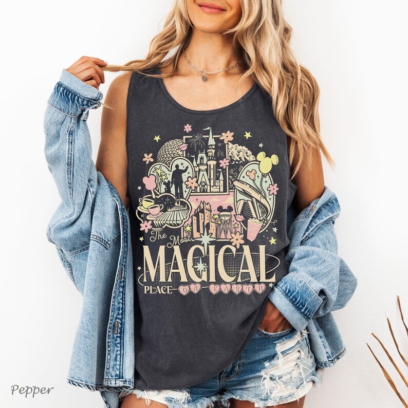 May include: A charcoal grey sleeveless tank top featuring a graphic print of a castle, a monorail, and various park icons in pastel pink, yellow, and white. The text reads The Most Magical Place on Earth, with small heart icons below the text.