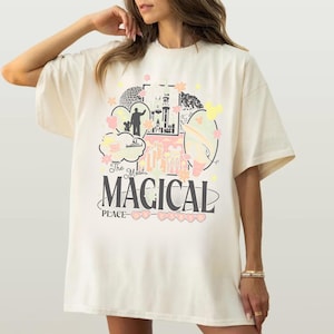 May include: An off-white short-sleeve t-shirt featuring a graphic print with a castle, park icons, and the text "The Most MAGICAL PLACE ON EARTH" in black lettering, accented with pastel pink, yellow, and green floral and character shapes.