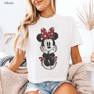 K&ouml;nnte beinhalten: Wei&szlig;es T-Shirt mit einem Used-Look-Motiv von Minnie Mouse. Die Zeichentrickfigur ist in Schwarzwei&szlig; mit einer roten Schleife mit Punkten dargestellt. Das Wort "White" ist in der oberen linken Ecke aufgedruckt.