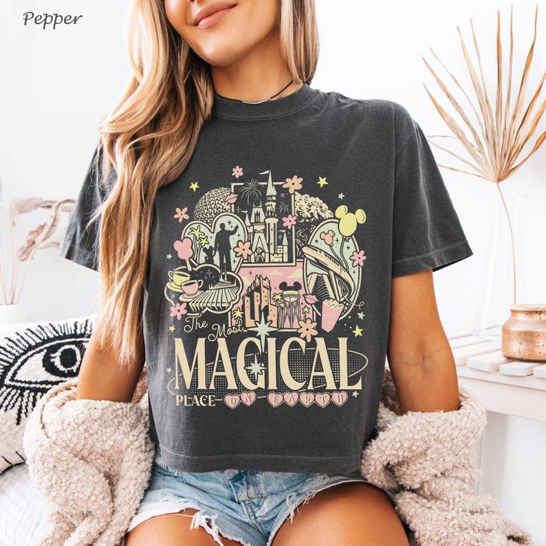 May include: A charcoal gray short-sleeve t-shirt featuring a graphic print with the text The Most Magical Place On Earth. The design includes illustrations of a castle, a monorail, teacups, and floral accents in shades of pink, yellow, and white.