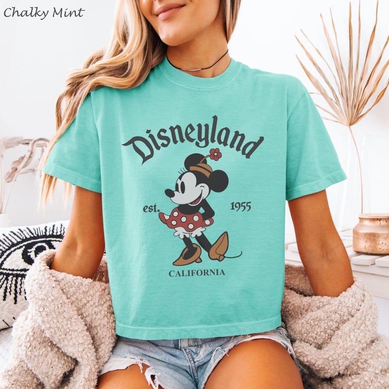 May include: A mint green t-shirt featuring a vintage Disneyland graphic. The design includes the word "Disneyland" in a script font, a Minnie Mouse illustration, and the text "est. 1955 CALIFORNIA".