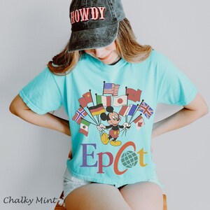 Puede incluir: Una camiseta azul claro con un dise&ntilde;o de Mickey Mouse sosteniendo banderas de varios pa&iacute;ses. La palabra "Epcot" est&aacute; impresa debajo del dise&ntilde;o. Se usa una gorra negra con la palabra "HOWDY".
