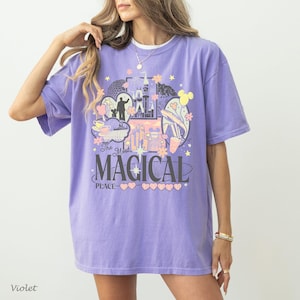 May include: A light purple short-sleeve t-shirt featuring a graphic print of a castle, a monorail, and a silhouette of a person with a mouse-eared figure. The text reads The Most Magical Place with small heart and star icons in pastel colors.
