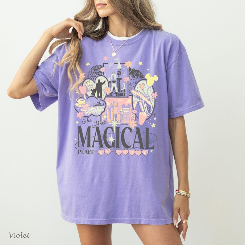 May include: A light purple short-sleeve t-shirt featuring a graphic print of a castle, a monorail, and a silhouette of a person with a mouse-eared figure. The text reads The Most Magical Place with small heart and star icons in pastel colors.