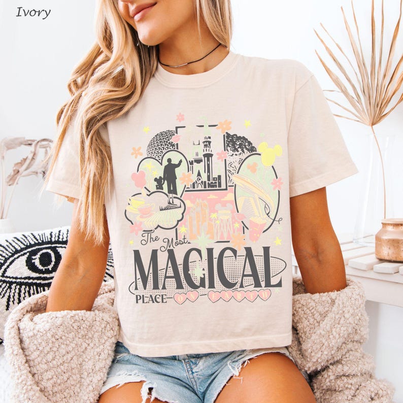 May include: An ivory t-shirt featuring a graphic print with the text "The Most MAGICAL PLACE" in dark gray. The design includes illustrations of a castle, a silhouette of a person with a mouse, teacups, and various pastel-colored floral shapes.