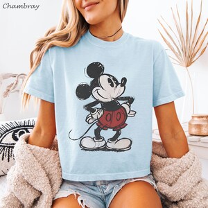 K&ouml;nnte beinhalten: Hellblaues T-Shirt mit einem verblichenen Mickey Mouse-Motiv. Die Zeichentrickfigur ist in Schwarz, Wei&szlig; und Rot dargestellt. Das Shirt hat kurze &Auml;rmel und einen Rundhalsausschnitt. Das Wort "Chambray" befindet sich oben links.