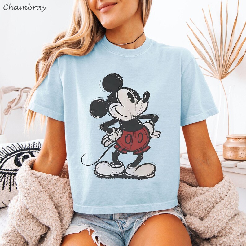K&ouml;nnte beinhalten: Hellblaues T-Shirt mit einem verblichenen Mickey Mouse-Motiv. Die Zeichentrickfigur ist in Schwarz, Wei&szlig; und Rot dargestellt. Das Shirt hat kurze &Auml;rmel und einen Rundhalsausschnitt. Das Wort "Chambray" befindet sich oben links.