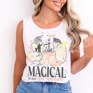 May include: A white sleeveless tank top featuring a colorful graphic print of a castle, a geodesic dome, and various park icons. The text reads "The Most MAGICAL PLACE ON EARTH" in black and pink lettering, accented by small flowers and stars.