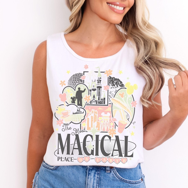 May include: A white sleeveless tank top featuring a colorful graphic print of a castle, a geodesic dome, and various park icons. The text reads "The Most MAGICAL PLACE ON EARTH" in black and pink lettering, accented by small flowers and stars.