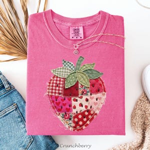 May include: A pink Comfort Colors t-shirt with a patchwork strawberry design. The strawberry is made of various patterned fabrics, including gingham, floral, and heart prints. The shirt also features a small silver necklace and the word "Crunchberry".