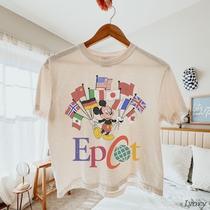 Puede incluir: Camiseta de color marfil con Mickey Mouse sosteniendo banderas de varios pa&iacute;ses. La palabra "Epcot" est&aacute; impresa en letras coloridas debajo del personaje, con un globo terr&aacute;queo. La camiseta est&aacute; colgada en una percha de madera.