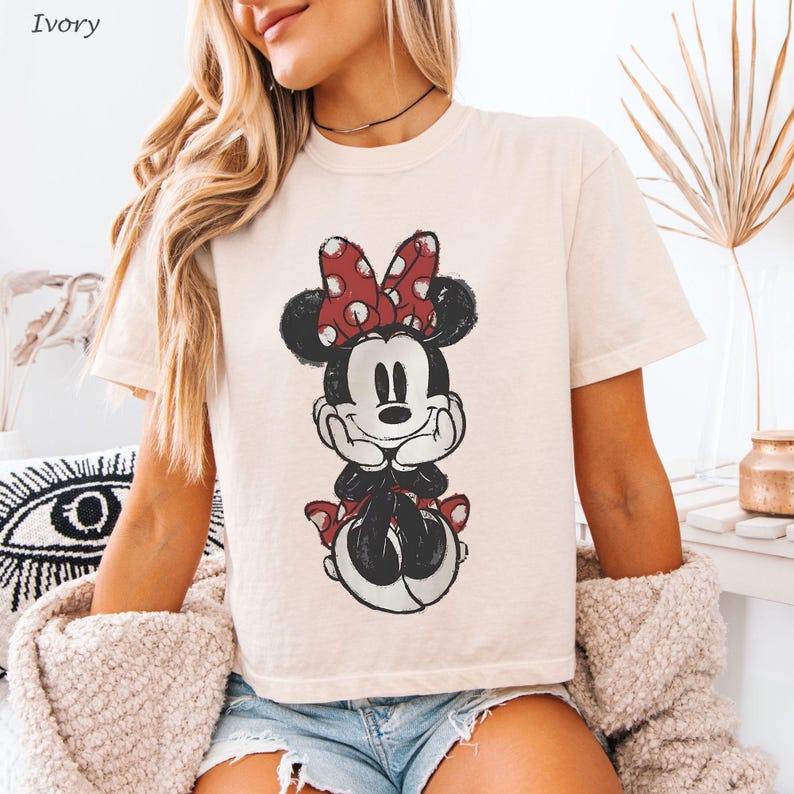 K&ouml;nnte beinhalten: Elfenbeinfarbenes T-Shirt mit einem verwaschenen Minnie Mouse-Motiv. Die Cartoonfigur ist in Schwarz, Rot und Wei&szlig; gehalten und tr&auml;gt eine rote Schleife mit Punkten. Das Oberteil hat kurze &Auml;rmel und einen Rundhalsausschnitt.