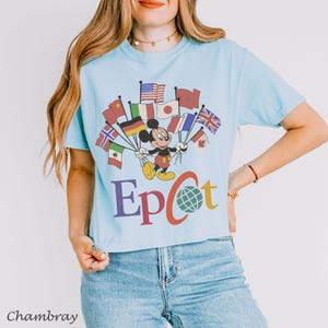 Puede incluir: Camiseta azul claro con Mickey Mouse sosteniendo banderas de todo el mundo. La palabra "Epcot" est&aacute; impresa en morado, rojo y verde. La camiseta est&aacute; hecha de un material suave.