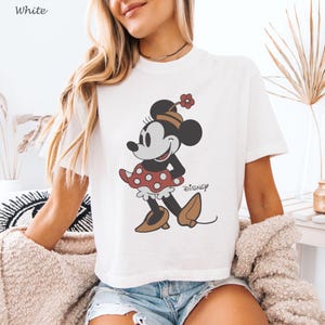 以下が含まれることがあります： ミニーマウスのグラフィックが特徴の白いTシャツ。漫画のキャラクターは、赤い水玉模様のスカート、茶色の靴、花の帽子で描かれています。「ディズニー」の文字が下に印刷されています。