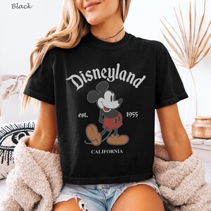 May include: Black t-shirt featuring a vintage Disneyland graphic. The design includes Mickey Mouse, the word "Disneyland", the year "1955", and the word "CALIFORNIA". The shirt has short sleeves and a crew neck.