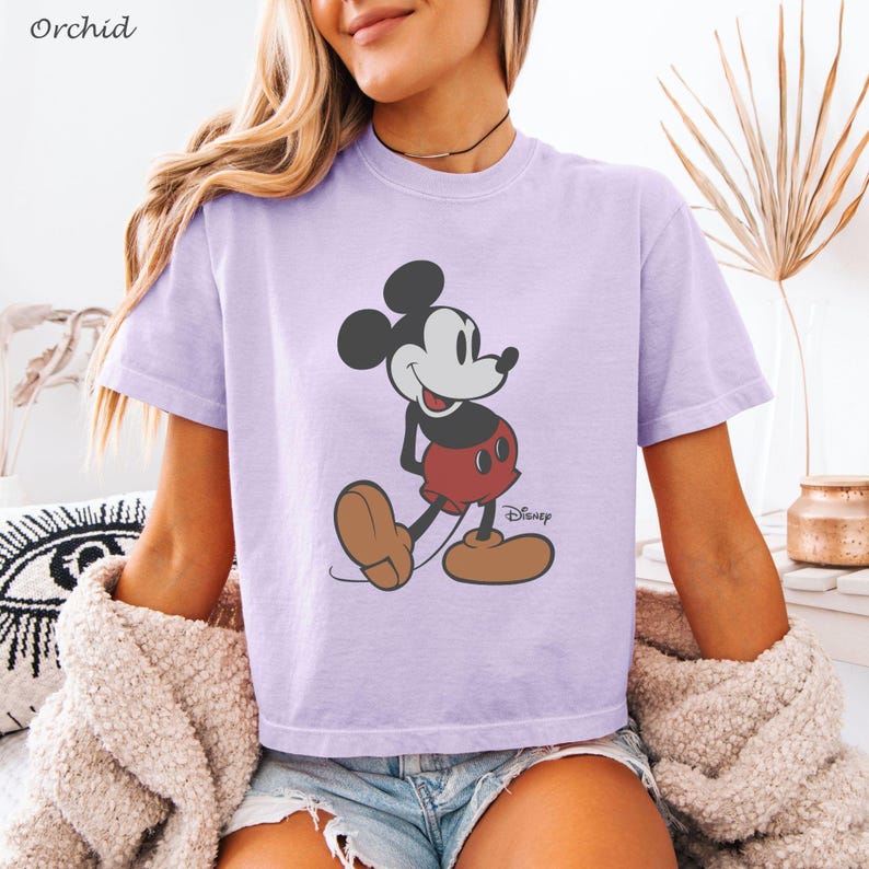 以下が含まれることがあります： ミッキーマウスが歩いているイラストが描かれた、薄紫色のTシャツ。ミッキーは、象徴的な赤いパンツ、茶色の靴、黒い手袋を着用しています。ディズニーのロゴが見えます。「Orchid」の文字が左上にあります。