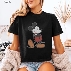 以下が含まれることがあります： ミッキーマウスが歩いている姿のグラフィックがプリントされた黒いTシャツ。ミッキーマウスは灰色の顔、黒い耳、赤いズボン、茶色の靴をしています。ディズニーのロゴが右下に表示されています。
