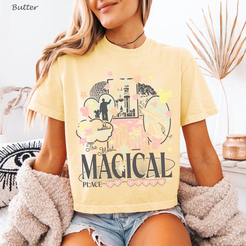 May include: A butter yellow short-sleeve t-shirt featuring a graphic print with the text "The Most MAGICAL PLACE" and illustrations of a castle, a silhouette of a person with a mouse, and various small star and flower shapes in pastel colors.