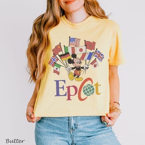 Puede incluir: Camiseta amarilla clara con Mickey Mouse sosteniendo banderas de todo el mundo. La palabra "Epcot" est&aacute; impresa debajo en un estilo retro. La camiseta es de color amarillo claro.