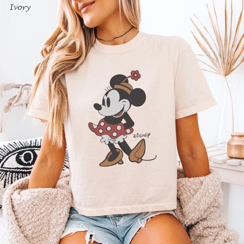 以下が含まれることがあります： ミニーマウスのグラフィックが特徴のアイボリー色のTシャツ。漫画のキャラクターは、赤い水玉模様のスカート、茶色の靴、花が付いたお揃いの帽子で描かれています。ディズニーのロゴが下に印刷されています。