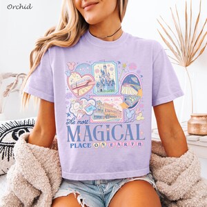 May include: A light purple t-shirt with a colorful graphic featuring various theme park icons and the text "The most MAGICAL PLACE ON EARTH". The shirt has short sleeves and a crew neck.
