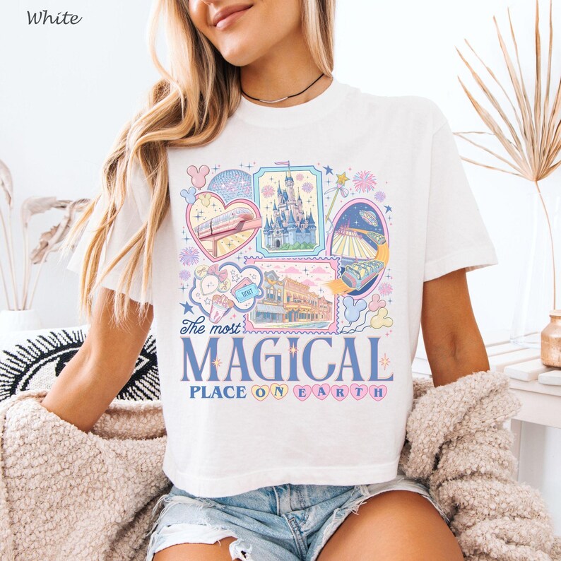 May include: White t-shirt with a colorful graphic design featuring various theme park attractions. The design includes a monorail, a castle, and the text "The most MAGICAL PLACE ON EARTH."
