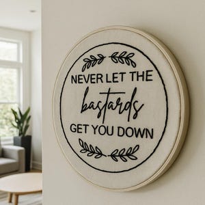 May include: Round, framed embroidery hoop with the text "NEVER LET THE bastards GET YOU DOWN" in black thread. The text is centered on a cream-colored fabric, surrounded by a black circular border and decorative leaves.