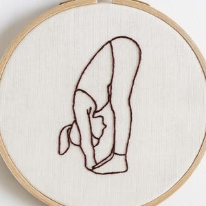 Puede incluir: Bastidor de bordado con un diseño minimalista de una persona en una postura de yoga de flexión hacia adelante. La obra de arte está cosida con hilo rojo oscuro sobre tela crema, dentro de un aro de madera natural.