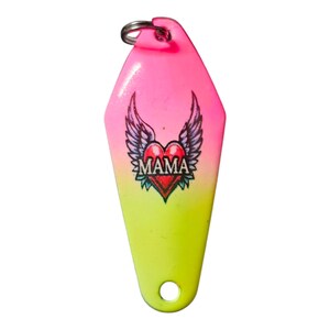 Könnte beinhalten: Rosa-gelber Angelköder mit Herz- und Flügeldesign. Das Herz trägt die Aufschrift "MAMA".