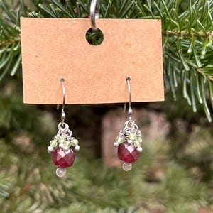 May include: A pair of dangle earrings with a silver hook and a cluster of small, light green beads. The earrings have a faceted, deep red gemstone and a small silver bead at the bottom. The earrings are displayed on a brown tag.