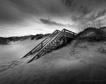 May include: A wooden staircase leading up a sand dune to a wooden walkway. The staircase is made of weathered wood and has a handrail on both sides. The sand dune is covered in grass and the sky is cloudy.