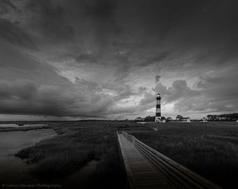 May include: A black and white photograph of a lighthouse on a cloudy day. A wooden walkway leads to the lighthouse, which is located on a grassy island. The sky is dark and stormy, but the lighthouse is lit up.