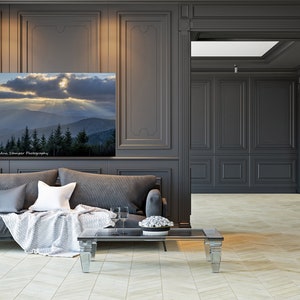May include: A gray sofa with pillows and a throw blanket sits in a room with dark gray walls and wood paneling. A framed photograph of a mountain landscape with a dramatic sky hangs on the wall above the sofa. A glass coffee table sits in front of the sofa.
