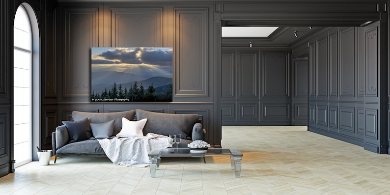 May include: A gray sofa with pillows and a throw blanket sits in a room with dark gray walls and wood paneling. A framed photograph of a mountain landscape with a dramatic sky hangs on the wall above the sofa. A glass coffee table sits in front of the sofa.