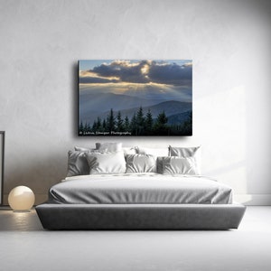 May include: A photograph of a mountain range with a dramatic sky. The sun is shining through the clouds, creating a ray of light. The mountains are covered in trees. The image is printed on canvas and framed.