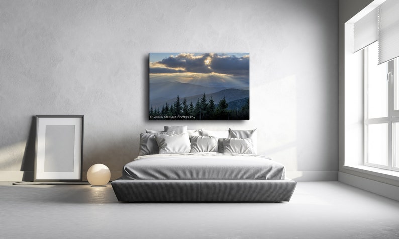 May include: A photograph of a mountain range with a dramatic sky. The sun is shining through the clouds, creating a ray of light. The mountains are covered in trees. The image is printed on canvas and framed.