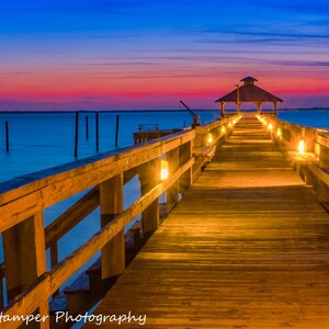 May include: A wooden pier extends out over the water at dusk. The pier is lit with string lights and leads to a gazebo at the end. The sky is a vibrant mix of pink, blue, and purple.