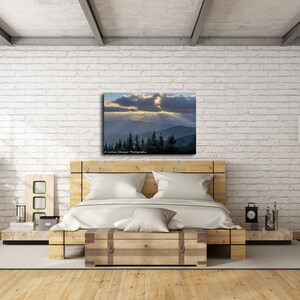May include: A bedroom interior with a white brick wall, a wooden bed frame, and a canvas print of a mountain landscape with a cloudy sky and sunbeams. The print is above the bed and is framed in a wooden frame. There is a wooden floor, a wooden side table, and a floor lamp with a white shade.