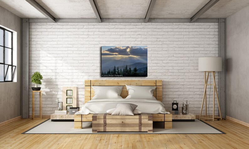 May include: A bedroom interior with a white brick wall, a wooden bed frame, and a canvas print of a mountain landscape with a cloudy sky and sunbeams. The print is above the bed and is framed in a wooden frame. There is a wooden floor, a wooden side table, and a floor lamp with a white shade.
