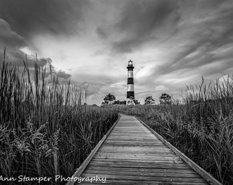 May include: A black and white photograph of a wooden walkway leading to a lighthouse on a cloudy day. The lighthouse is tall and striped with black and white bands. The walkway is surrounded by tall grass.