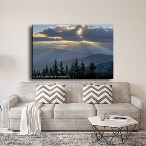 May include: A photograph of a mountain range with a cloudy sky and sunbeams shining through the clouds. The mountains are covered in trees and the sky is a light blue color. The image is taken from a high vantage point and the mountains are in the distance.