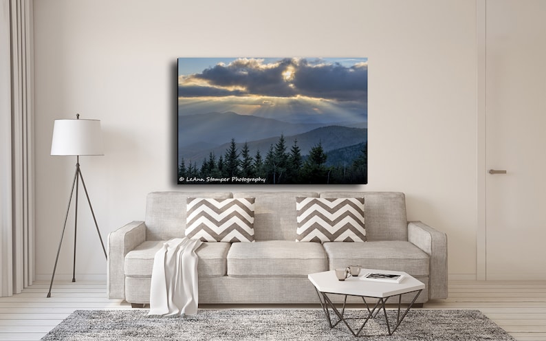 May include: A photograph of a mountain range with a cloudy sky and sunbeams shining through the clouds. The mountains are covered in trees and the sky is a light blue color. The image is taken from a high vantage point and the mountains are in the distance.