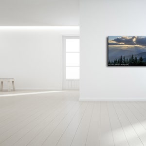 May include: A photograph of a mountain range with a sunbeam shining through the clouds. The mountain range is covered in trees and the sky is a light blue color.
